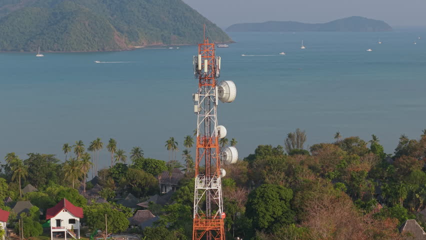  Aerial 4K footage of a telecommunication tower surrounded by lush greenery and a coastal landscape.