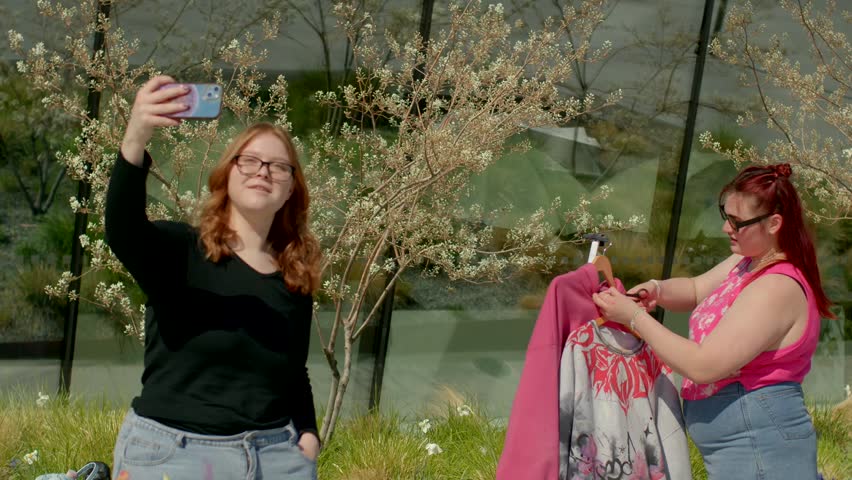 Two women delighting in a beautiful sunny day outdoors, happily posing for selfies while showcasing their trendy clothing