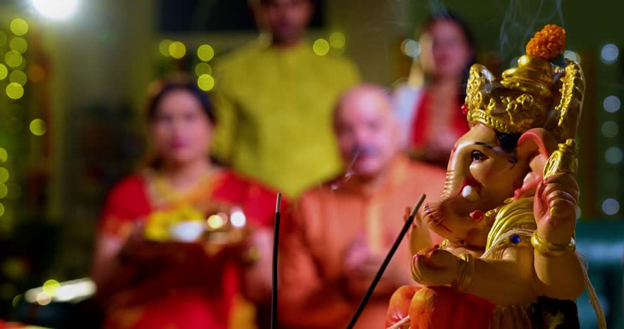Focus on Lord Ganesha idol during Ganesh Chaturthi as Indian joint family with senior parents, son, daughter-in-law offer prayers with laddu plate and puja thali indoors in decorated festive home