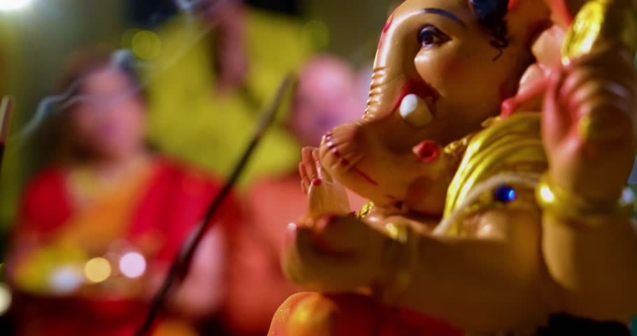 Focus on Lord Ganesha idol during Ganesh Chaturthi as Indian joint family with senior parents, son, daughter-in-law offer prayers with laddu plate and puja thali indoors in decorated festive home