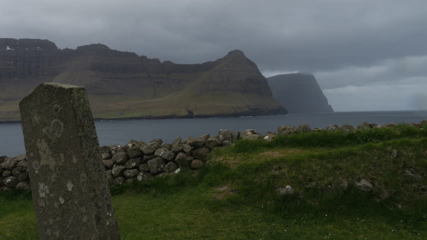 Ancient stone stands in a grassy field, overlooking a stone wall and the coastline of the faroe islands under a cloudy sky
