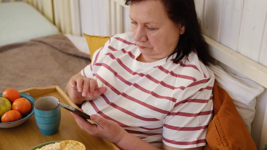 Senior woman having breakfast sitting in bed, drinking tea, enjoying her morning at home. Old lady eating from tray and using phone. Elderly female in pajamas relaxing with cup of coffee in a bedroom.