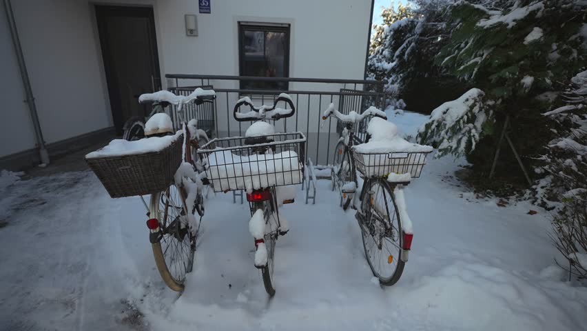 Three snow-covered bicycles parked outside a small apartment house in a German forest. Baskets full of snow, peaceful winter scene, rural lifestyle, quiet snowy day in suburban Europe. 