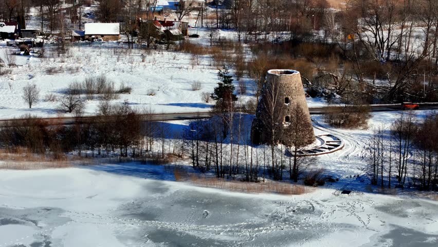 An abandoned stone tower stands near a partially frozen lake in a snow-covered countryside, framed by bare trees and quiet rural scenery during the winter season from an aerial angle.