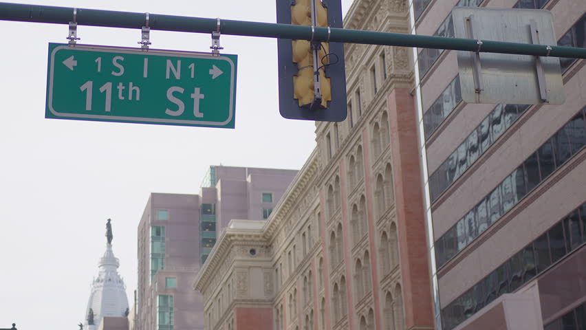Slow-motion clip of the 11th St sign gently swaying in the wind on a traffic light, with Philadelphia’s Capitol dome, the Ben Franklin statue, and nearby buildings visible in the scenic background.