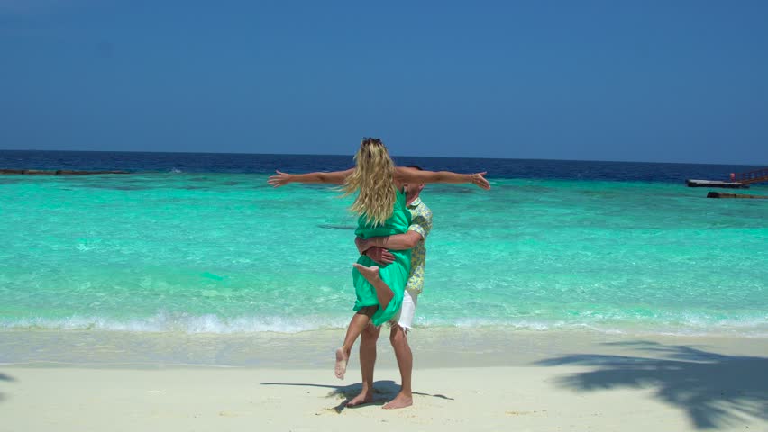 A couple spins around joyfully on a beach in the Maldives, with turquoise water and sandy shore in the background.