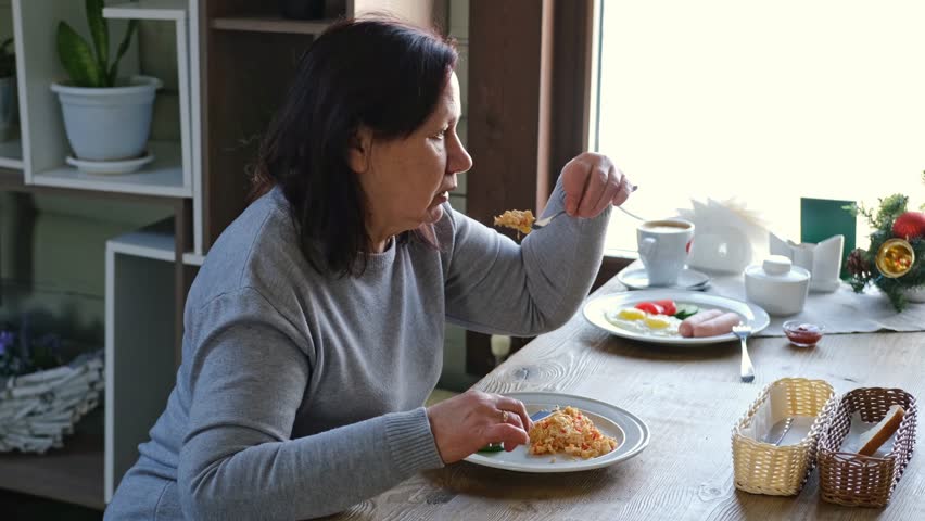 Happy senior woman eating a scramble on a white plate with vegetables. Elderly lady using knife and fork to cutting meal, has a breakfast. Balanced diet, cooking, culinary. Food concept. Old woman