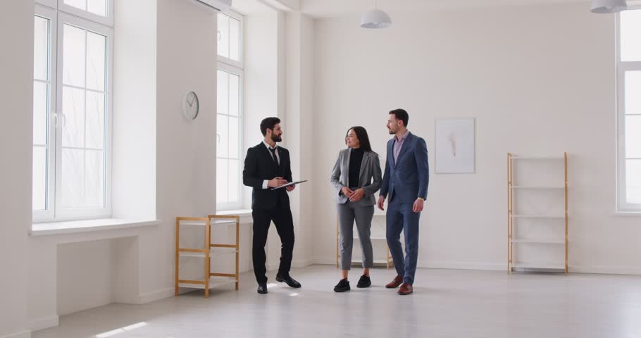 Real estate agent showing property or empty office room to woman and man, giving a tour about, telling about advantages of buying this dwelling. Realtor showing potential buyes apartment