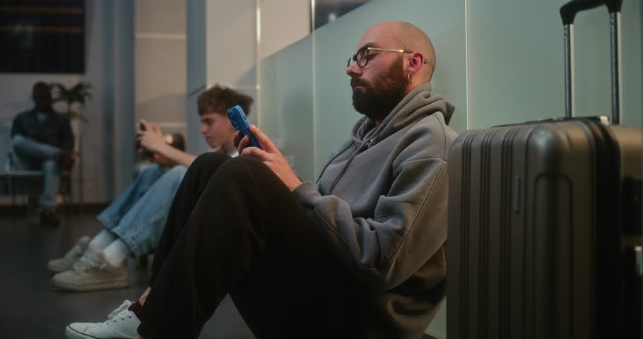 Diverse People and Passengers in Crowded International Airport Terminal: Tired Man with Luggage Sitting on the Floor in Boarding Lounge of Airline Hub, Using Phone, Waiting for Delayed Airplane Flight