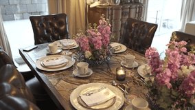 Sumptuous dining table is set with white china cups on gold‑rimmed chargers, polished cutlery and linen napkins around a centerpiece of pink stock blooms and a lit votive candle, slow motion - Powered by Shutterstock - Get 15% off with code: PIKWIZARD15
