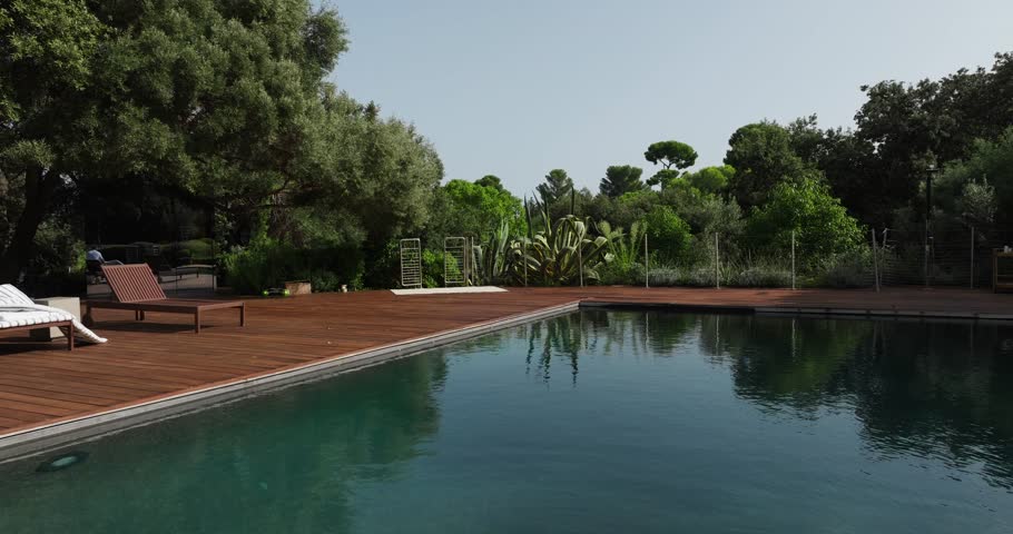Cinematic view of an elegant swimming pool surrounded by a wooden deck, lush trees, and cacti, capturing upscale outdoor living in a luxurious South of France real estate setting.