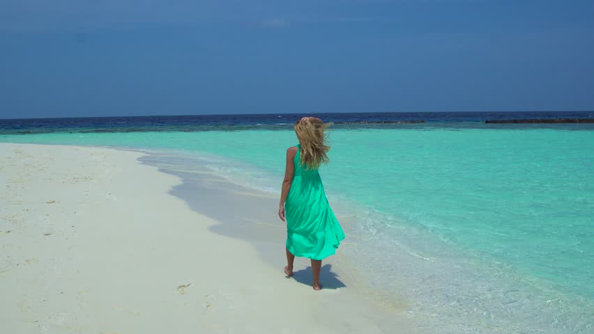 Woman walks along a sandy beach in the Maldives, ocean waves gently lapping at her feet.