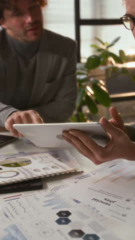 Vertical view of two young businessmen reviewing statistical data on digital tablet and discussing new strategy at office desk