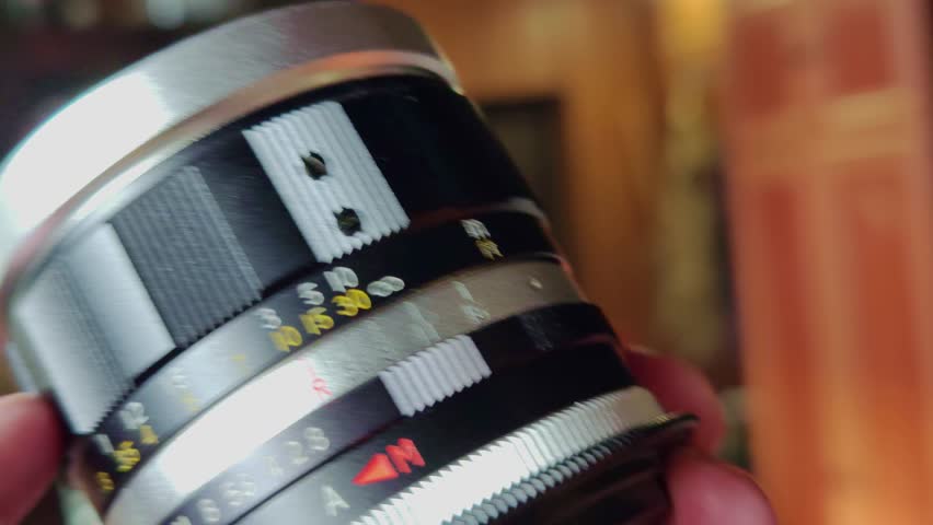 A close-up shot of a hand carefully holding a vintage camera lens. The lens features intricate markings for focus distance and aperture settings, with a silver top ring and a black and white body