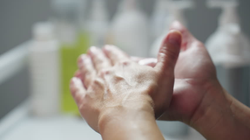 Facialist rubs clear massage gel between hands to evenly coat palms and fingers, preparing smooth hygienic product layer before applying to client face in bright spa treatment environment