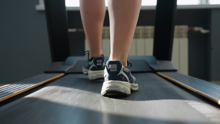 close up back view of person walking on treadmill belt under sunlight shining on shoes and legs in modern gym studio with exercise bike and elliptical trainer blurred behind