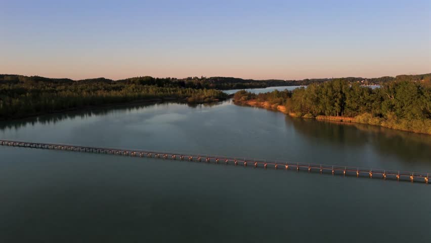 Drone footage showing a wooden vehicular causeway bridge connecting the west shore of Lake Woerthsee to Mausinsel, a private forested island in Bavaria, Germany. Peaceful and remote. 