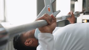 close up of man in white jacket pressing barbell bench press with headphones around neck, bright gym environment with windows and equipment in background, focus on upper body strength - Powered by Shutterstock - Get 15% off with code: PIKWIZARD15