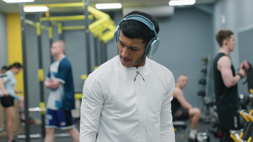 portrait view fitness enthusiast wearing over ear headphones and white zip top performing dumbbell lateral raise, focusing on form in busy gym environment with other athletes training in background
