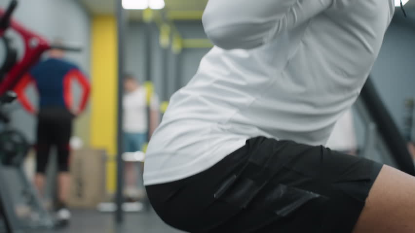 side view of student squatting with barbell under pressure in modern gym environment while other people train in background, wearing black shorts with reflective patch on leg