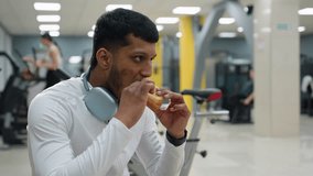 young man dressed in white activewear seated calmly in gym while chewing snack, wearing headphones around neck with blurred background of people exercising and gym equipment - Powered by Shutterstock - Get 15% off with code: PIKWIZARD15