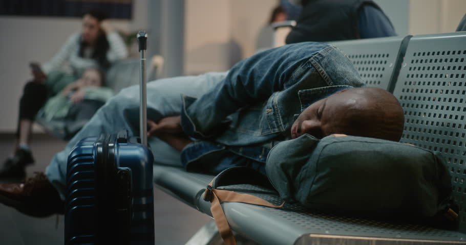 Crowded International Airport Terminal: Tired African American Tourist with Suitcase and Backpack Sleeping on the Seats in Boarding Lounge of Airline Hub. Diverse Passengers Waiting for Delayed Flight