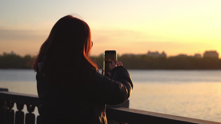 Young woman in a jacket is taking a photo or video of the sunrise or sunset by the river, sea, lake