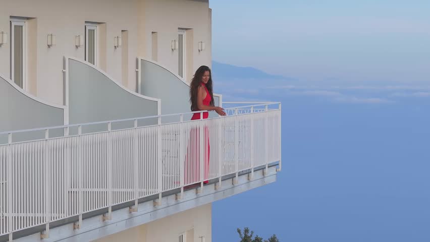 A woman in a red dress stands on a hotel balcony with white railings, overlooking the Amalfi coastline. Subtle motion shows her shifting posture.