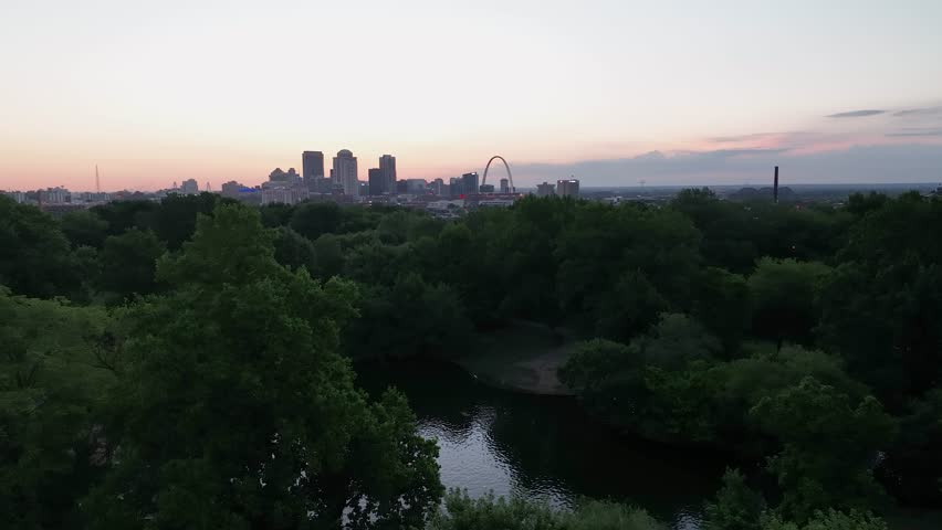 A cityscape view of Saint Louis, Missouri from a park with trees and a pond of water during sunrise.