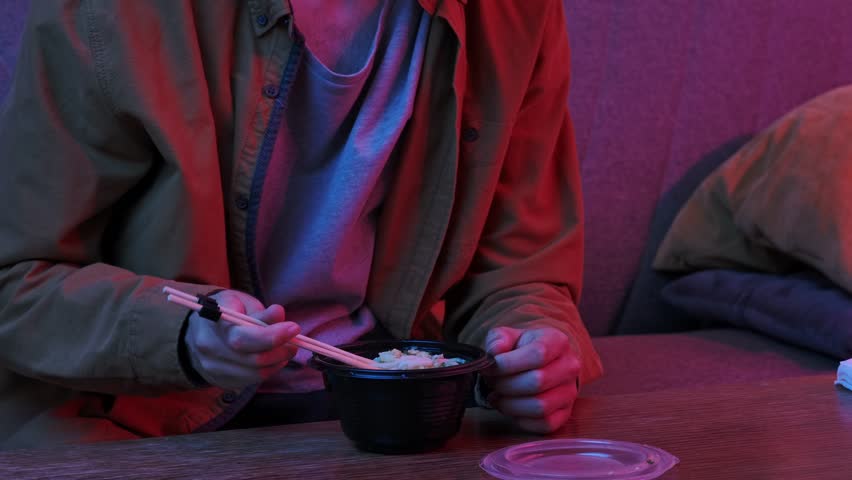 Enjoying asian fast food at home. Young man sitting at home in evening on the sofa, ordered delivery of tasty asian food, holding chopsticks and eating noodles soup from the bowl. Slow motion.
