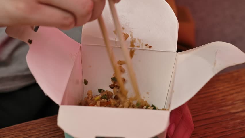 Enjoying asian fast food at home. Young man sitting at home in evening on the sofa, ordered delivery of tasty asian food, holding box, mixing and eating fried rice with chopsticks. Slow motion.