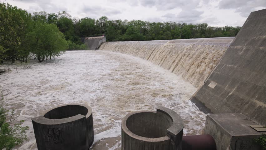 Slow motion wide landscape of a concrete dam holding and overflowing with water, waves, bubbles, minerals, and industrial equipment after recent rainfall, causing a strong stream and crashing liquid