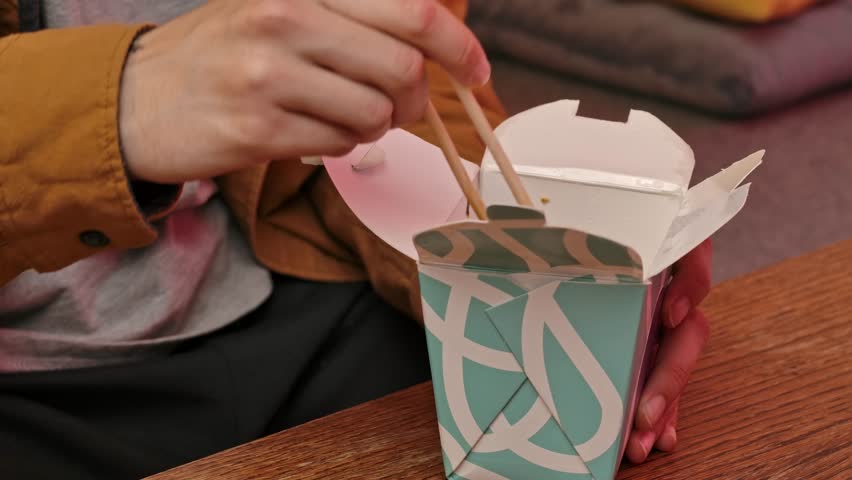 Enjoying asian fast food at home. Young man sitting at home in evening on the sofa, ordered delivery of tasty asian food, holding box, mixing and eating fried rice noodles with chopsticks. Slow motion