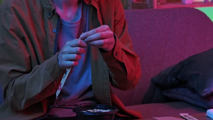 Enjoying asian fast food at home. Young man sitting at home in evening on the sofa, ordered delivery of tasty asian food, takes chopsticks prepares to eat cup noodles with miso soup in plastic bowl.