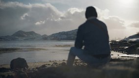 A man sits alone on a rocky shoreline, gazing at snow-covered mountains across the ocean as waves crash gently against the rocks - Powered by Shutterstock - Get 15% off with code: PIKWIZARD15