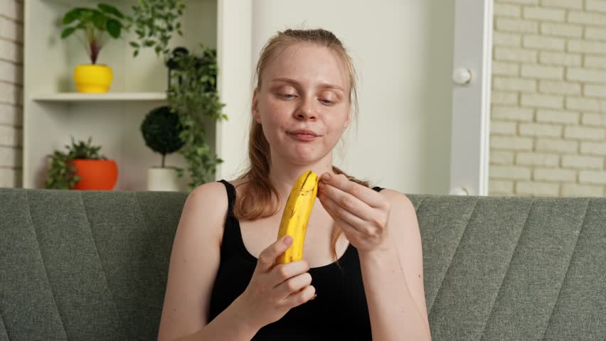 Healthy food advertisement concept. Young woman sitting on sofa at home and eating fresh vitamin fruit, peeling off and biting ripe banana, chewing and smiling, enjoying snack. Slow motion.
