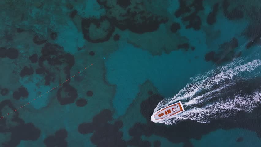 Aerial view of Capri, Italy, showing turquoise waters, underwater terrain, and boats. One boat in motion leaves a white wake across the sea.