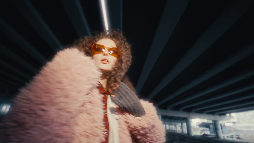 Stylish young woman with curly hair, wearing trendy sunglasses and pink fluffy fur coat, looking into camera and dancing energetically under urban bridge