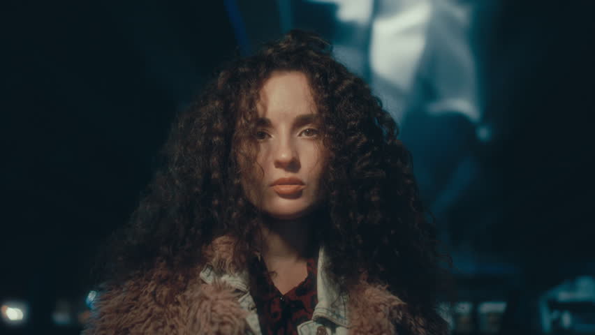 Young attractive woman with voluminous curly hair looking confidently into camera, standing in dark underpass illuminated by blue light at night. Video portrait