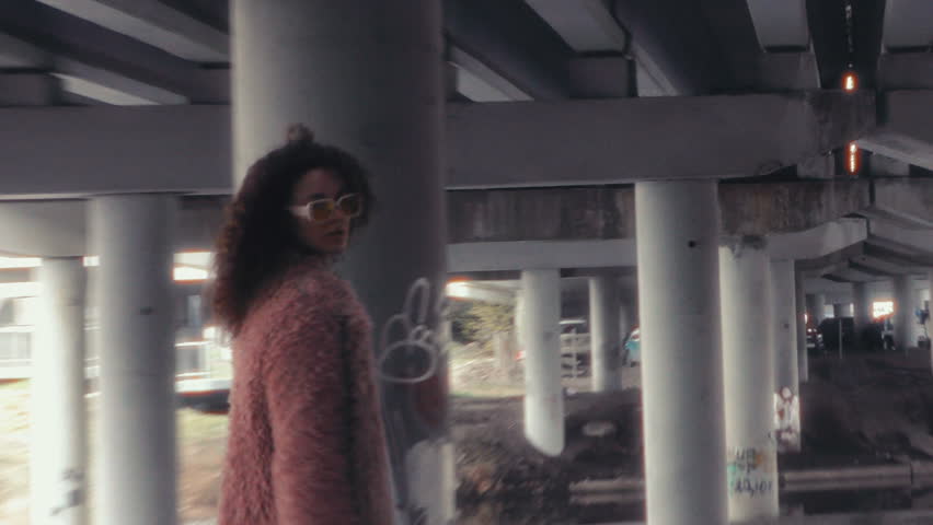 Stylish curly-haired girl in pink faux fur coat and sunglasses turning back and posing for camera as walking under highway overpass with concrete columns. Zoom handheld shot
