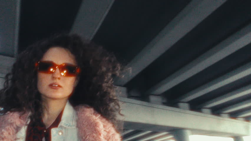 Young attractive woman with curly hair, wearing trendy fur coat and sunglasses, dancing and looking confidently into camera under concrete urban overpass. 360-degree shot