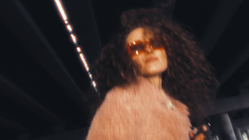 Young confident girl in pink faux fur coat and stylish sunglasses dancing for camera under highway concrete overpass in city. Zoom shaky camera shot