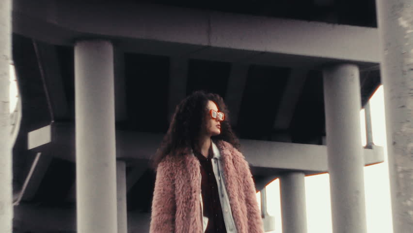 Confident girl in pink faux fur coat and sunglasses standing under massive concrete overpass, looking to side then turning to camera. Zoom shot, video portrait