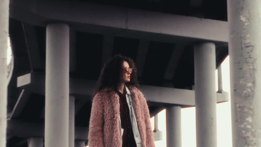 Young beautiful woman in trendy pink faux fur coat and sunglasses standing under urban concrete overpass, looking confidently to side then sharply turning to camera. Zoom shot, video portrait