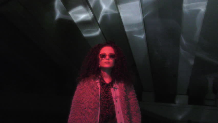 Young stylish woman in faux fur coat and sunglasses illuminated by red neon light, posing on camera under dark concrete overpass at night, Zoom shot, video portrait