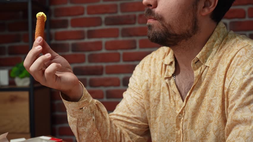 Junk food advertisement concept. Adult stylish bearded man sitting at table and holding grilled cheese stick with topping in hand, taking a bite and chewing, enjoying tasty fast food. Slow motion.