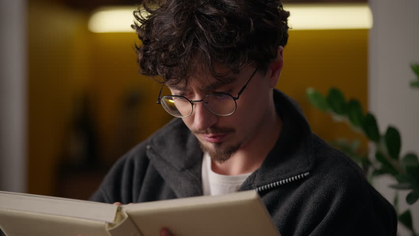 Pensive focused Caucasian guy hipster holding book notebook man reading interesting literature novel hobby leisure reader learn knowledge concentrated male putting on eyeglasses think at home library