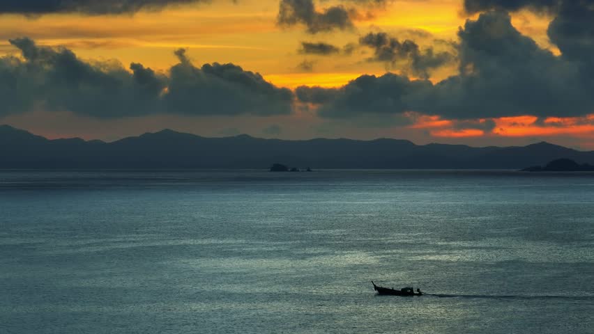 An aerial shot of a boat gliding across the ocean, with dark mountain silhouettes and a golden sunset peeking through stormy clouds, evoking a sense of peaceful solitude and nature’s power. Thailand.
