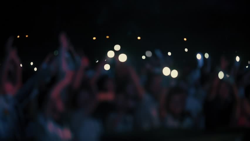 A stunning slow-motion footage of a large crowd at a night concert, waving hands and mobile phones with flashlights in the air. The audience follows the rhythm of the music, creating a magical and emo