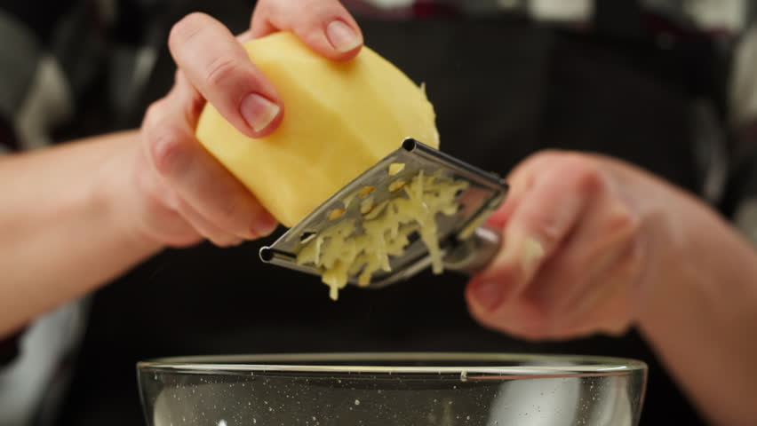 Woman hands with a grater grate the potatoes on the wooden board, chef cooking polish potato pancakes in the kitchen. Healthy eating and lifestyle. 